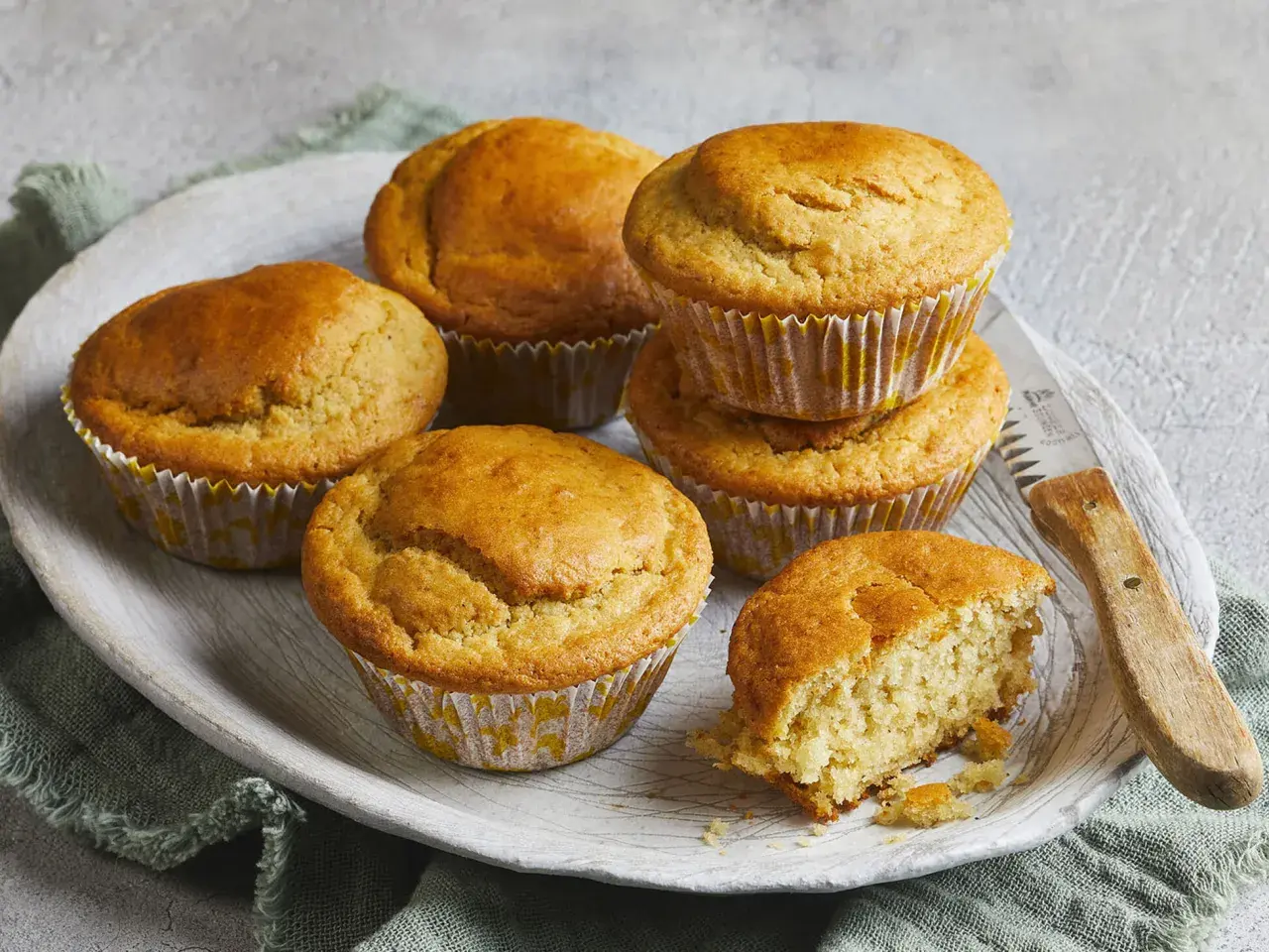 Goldbraune Buttermilch Muffins auf einem Holzteller, einer davon angebrochen, mit einem Messer daneben.