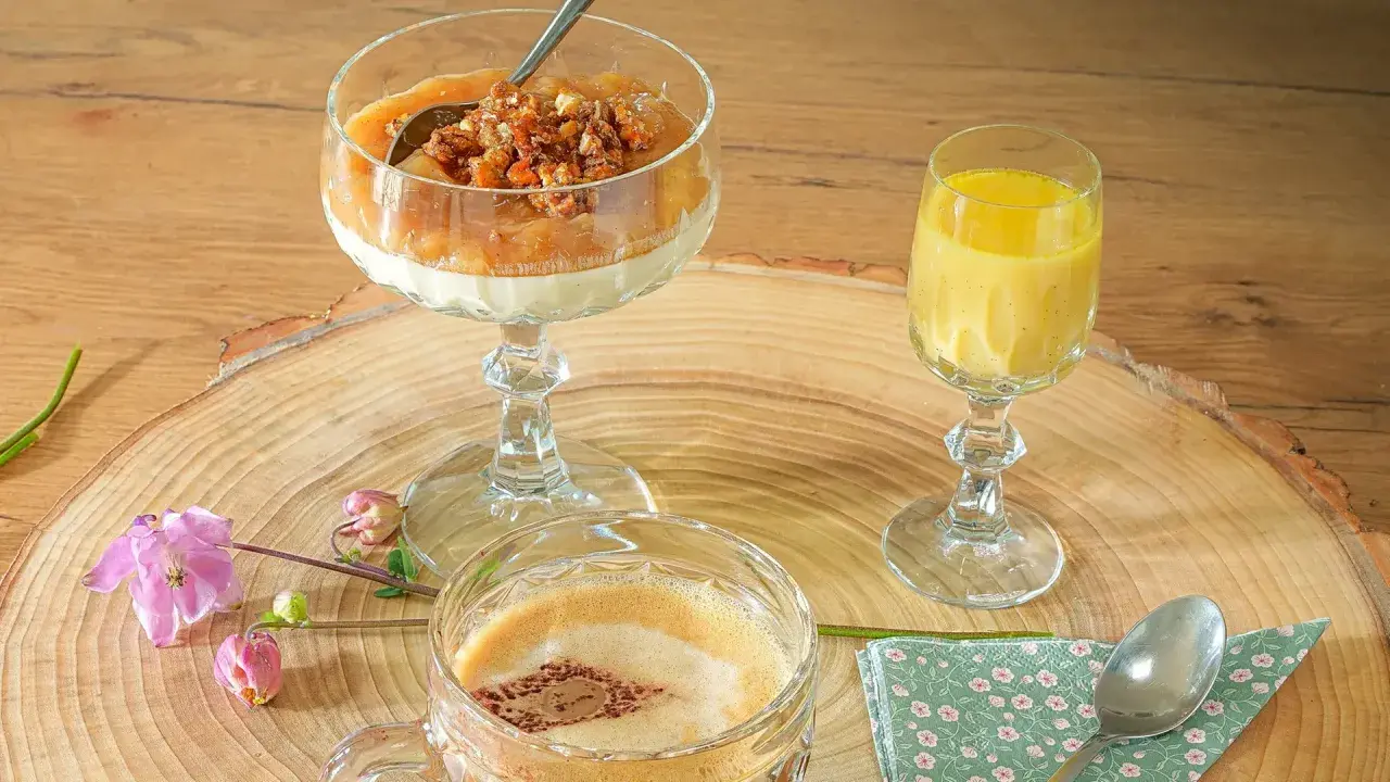 Ein Dessert im Glas mit Rhabarberkompott und Streuseln, daneben ein gelbes Getränk und ein Kaffee.