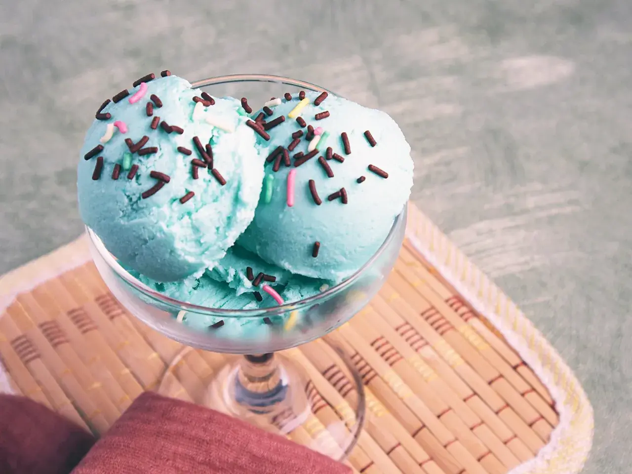 Zwei Kugeln blaues Eis mit bunten Streuseln in einem Glas auf einem Holzuntersetzer.