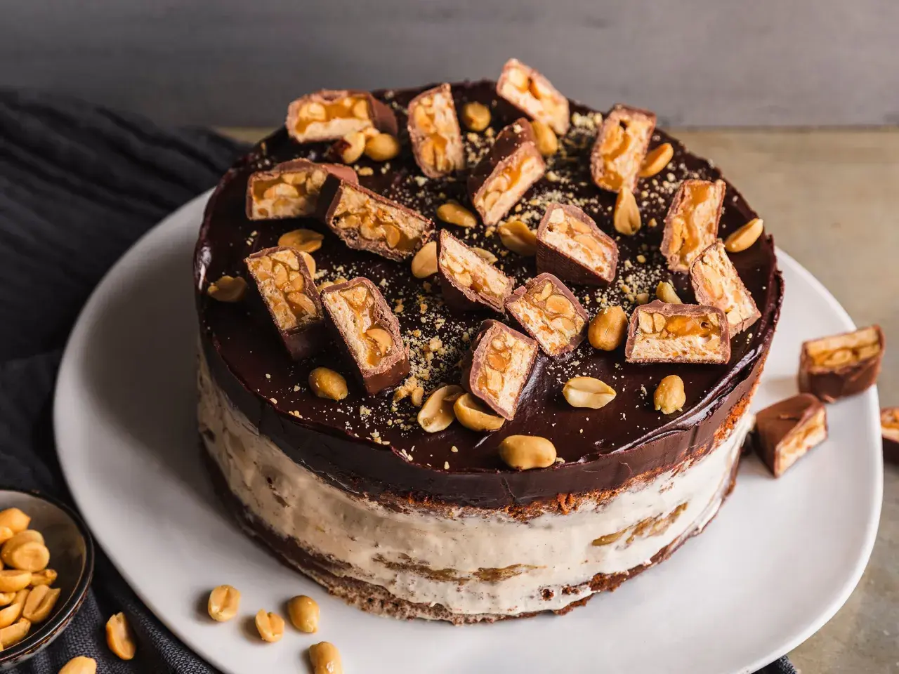 Eine köstliche Snickers Torte mit Schokoladenglasur, Erdnüssen und vielen Snickers-Stücken obendrauf.
