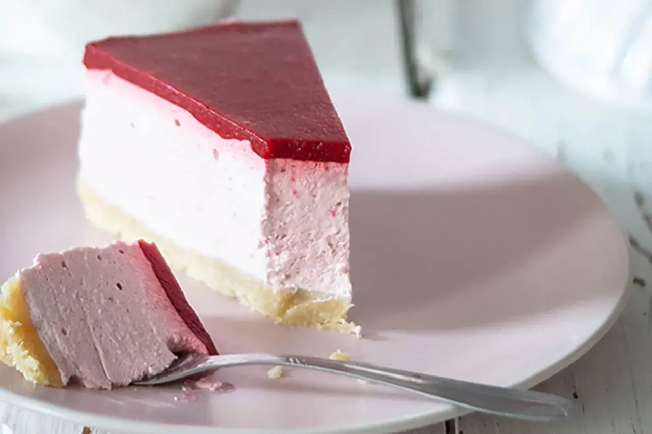 Ein Stück cremiger cheesecake ohne backen mit roter Fruchtschicht auf rosa Teller, daneben ein Gabel mit einem Stück Kuchen.
