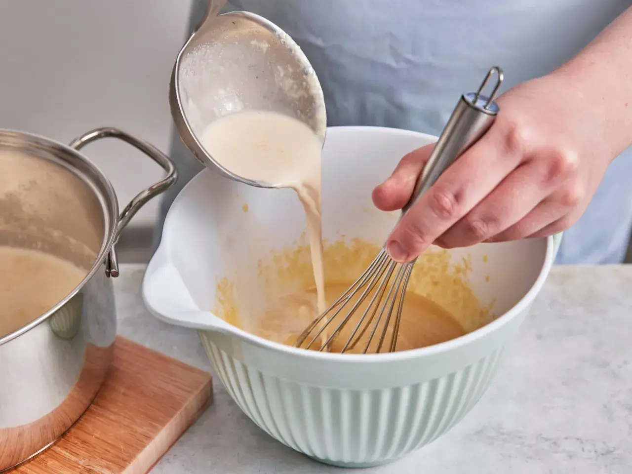 Eine cremige Masse wird aus einem Topf in eine Schüssel gegossen, während mit einem Schneebesen gerührt wird. Perfekt für selbstgemachtes Zimteis aus der Eismaschine.