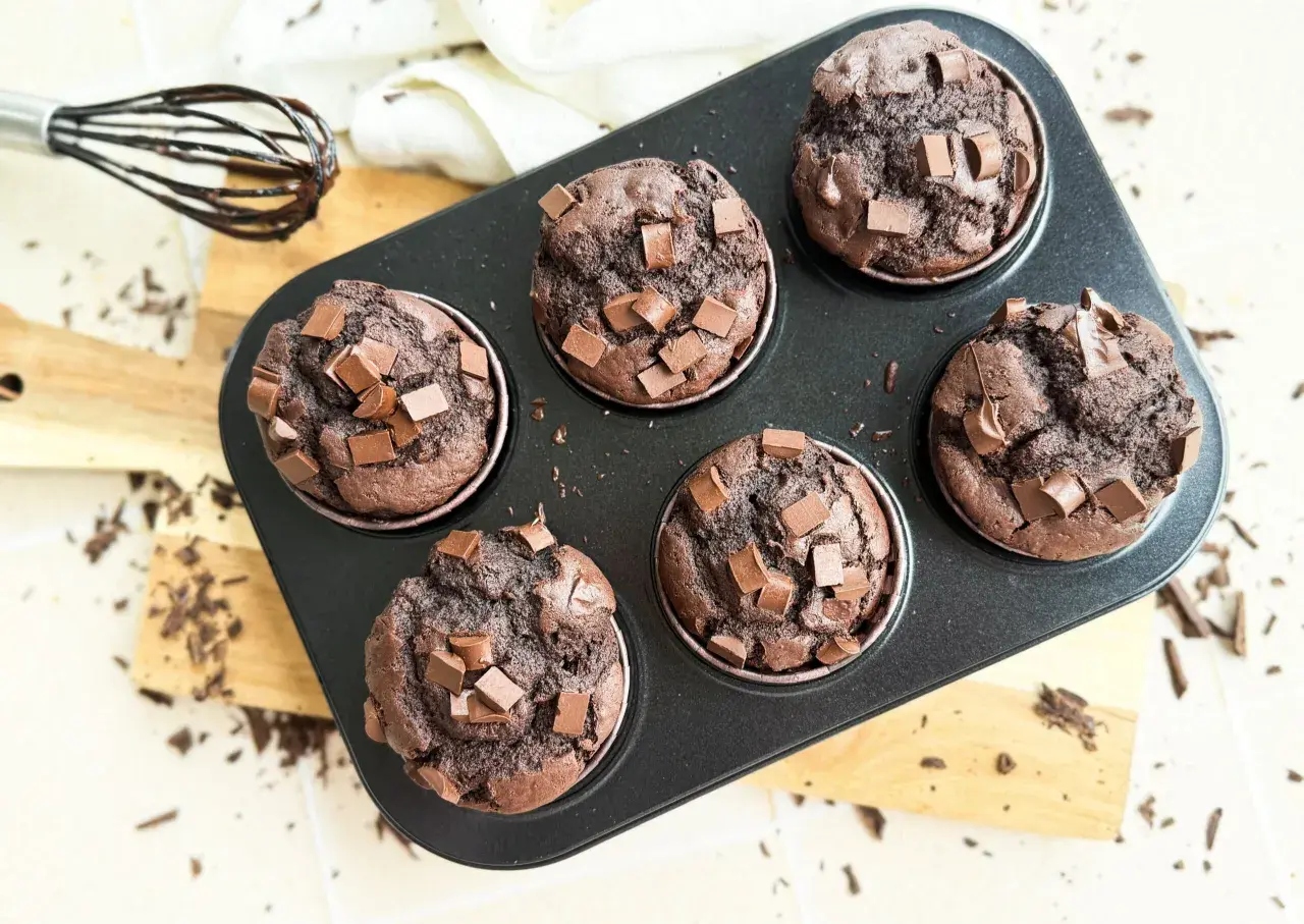 Sechs saftige, vegane Schokomuffins mit Schokostückchen in einer Muffinform. Ein Schneebesen mit Schokospuren liegt daneben.