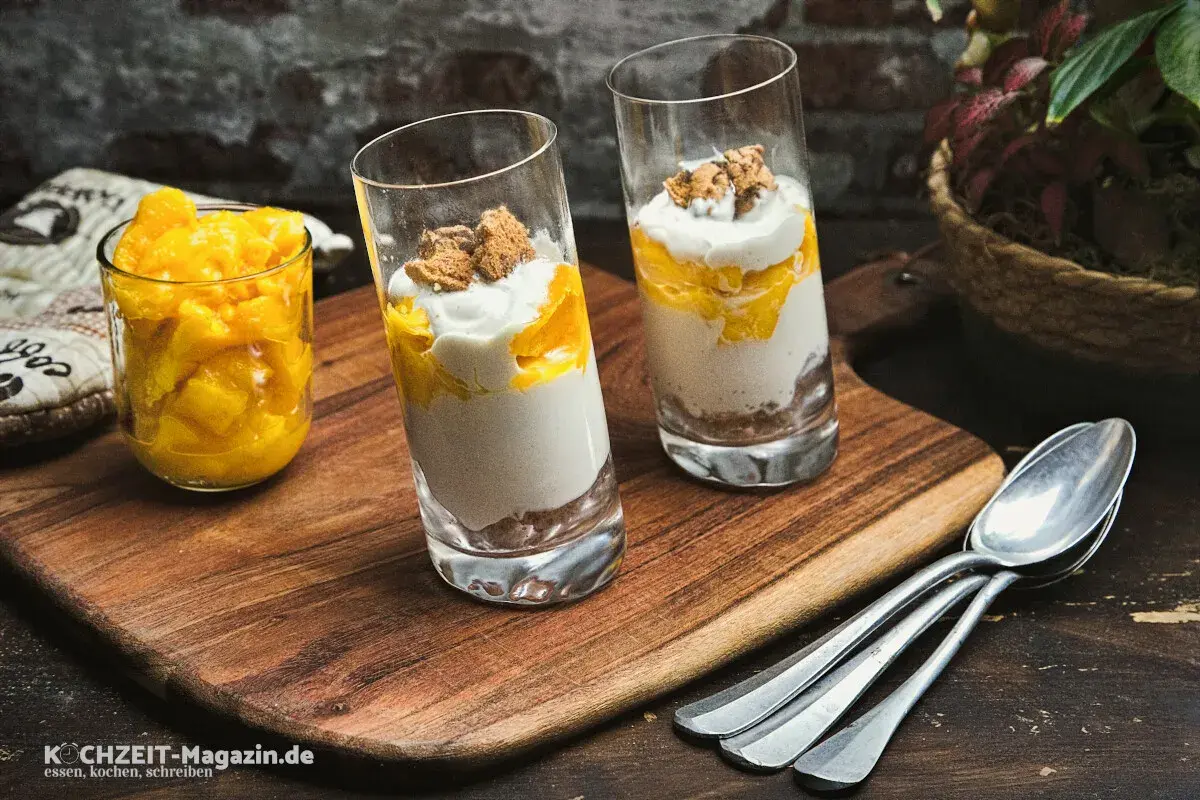 Zwei Gläser mit einem cremigen Mango-Dessert, geschichtet mit zerbröselten Keksen und garniert mit Mango-Stücken. Daneben ein Glas voll frischer Mango-Würfel.