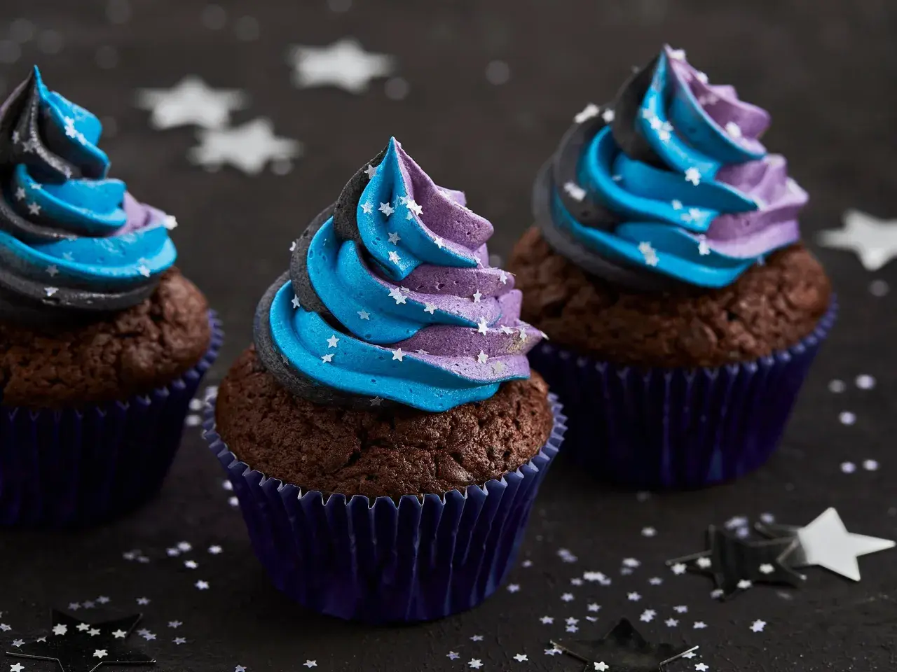Drei Schoko-Cupcakes mit blau-lila Sternen-Frosting, inspiriert von einem Galaxie-Cupcake-Rezept.