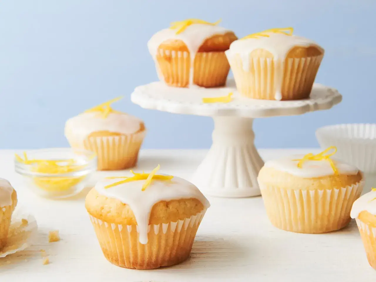 Saftige vegane Zitronenmuffins mit Zuckerguss und Zitronenschale verziert, auf einem weißen Kuchenständer und Tisch.