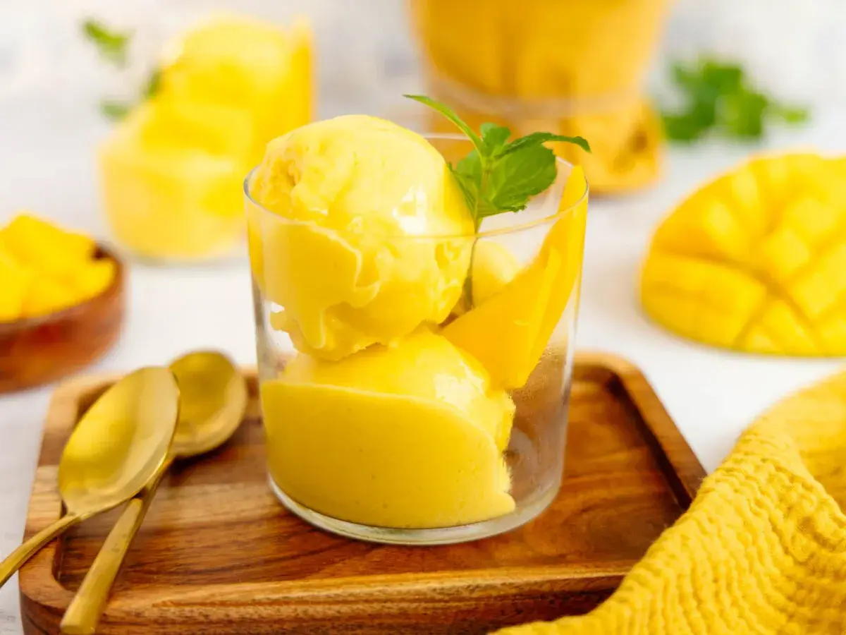 Erfrischendes Mango Eis in einem Glas, garniert mit Minze und Mangostücken. Daneben liegen goldene Löffel auf einem Holztablett.