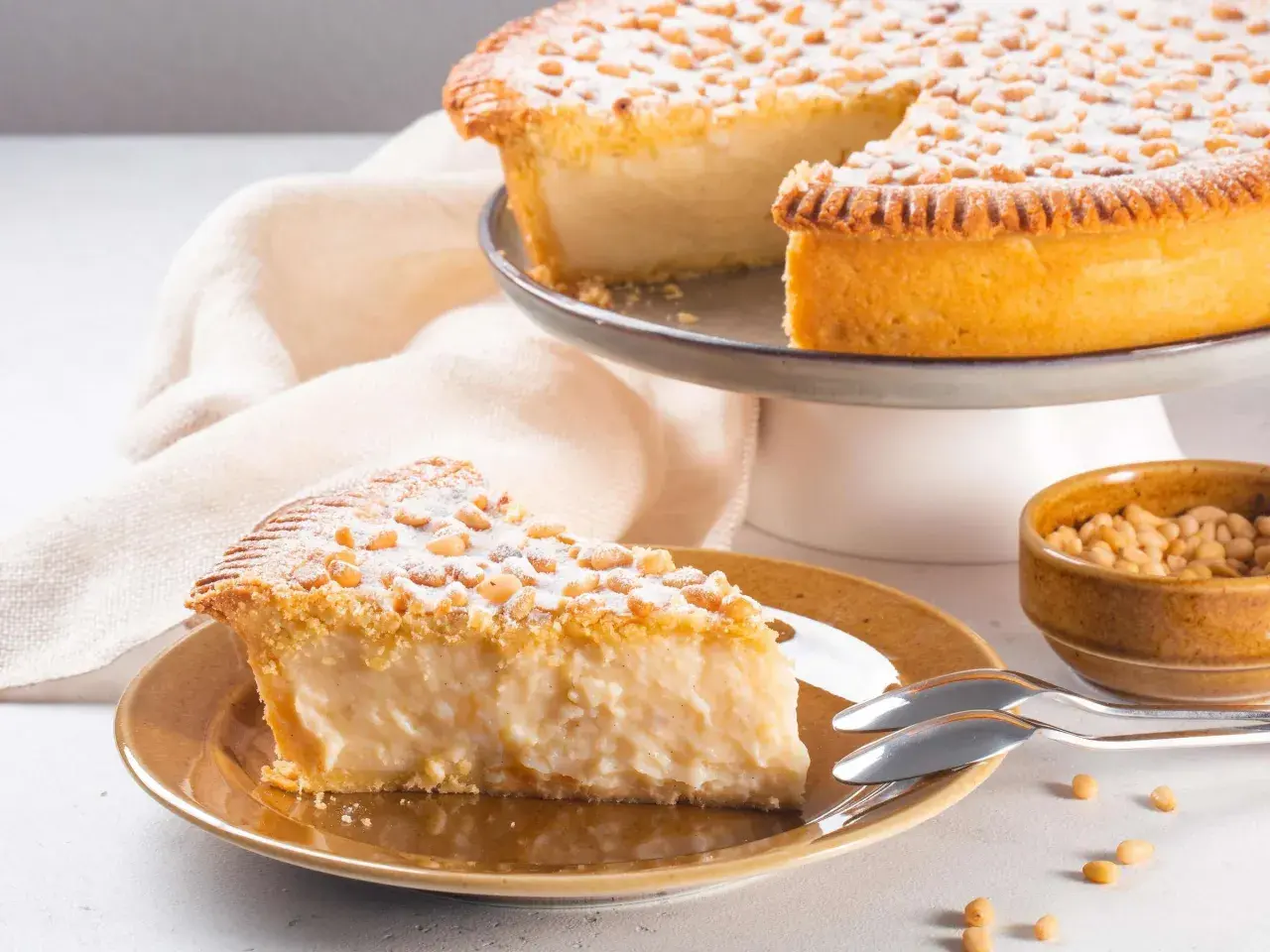 Ein Stück Torta della Nonna, bestäubt mit Puderzucker und Pinienkernen, auf einem Teller neben einer Schüssel voller Pinienkerne.