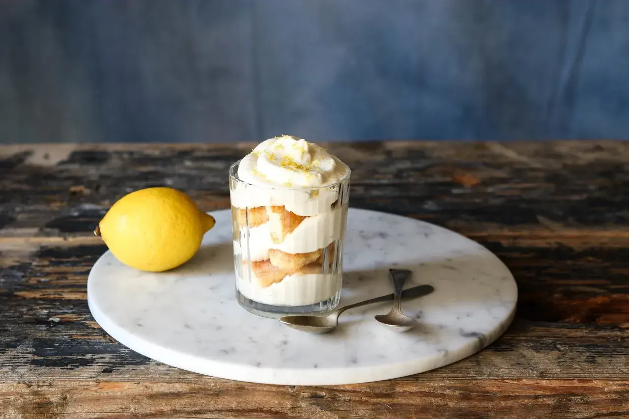 Ein Glas mit geschichtetem Zitronen Tiramisu, garniert mit Zitronenschale. Daneben liegt eine Zitrone.