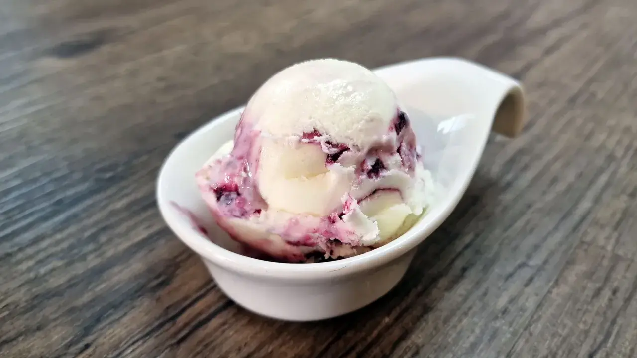 Cremiges Cheesecake Eis mit fruchtigen Beerenwirbeln in einer weißen Schale auf Holztisch.