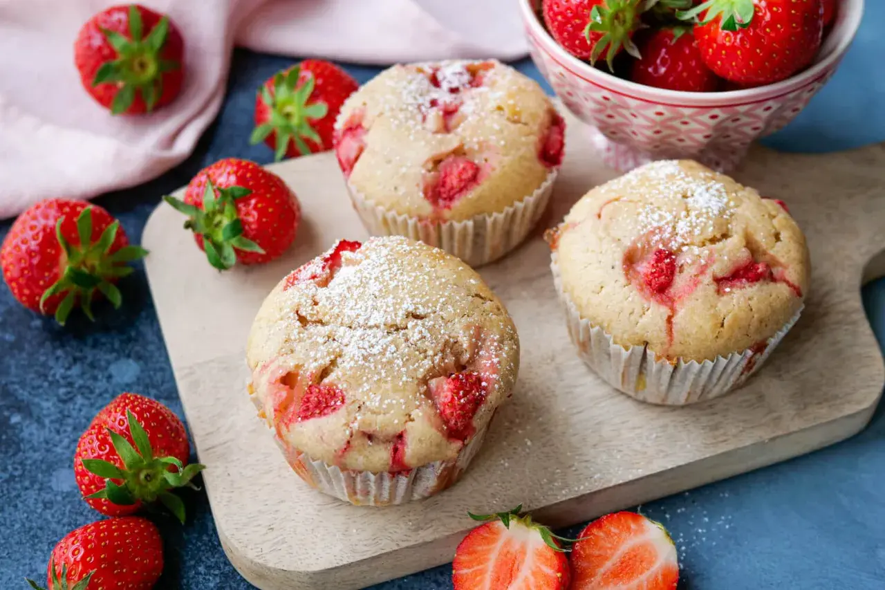 Saftige Erdbeer Muffins, bestäubt mit Puderzucker, liegen auf einem Holzbrett. Frische Erdbeeren umgeben die Leckereien.
