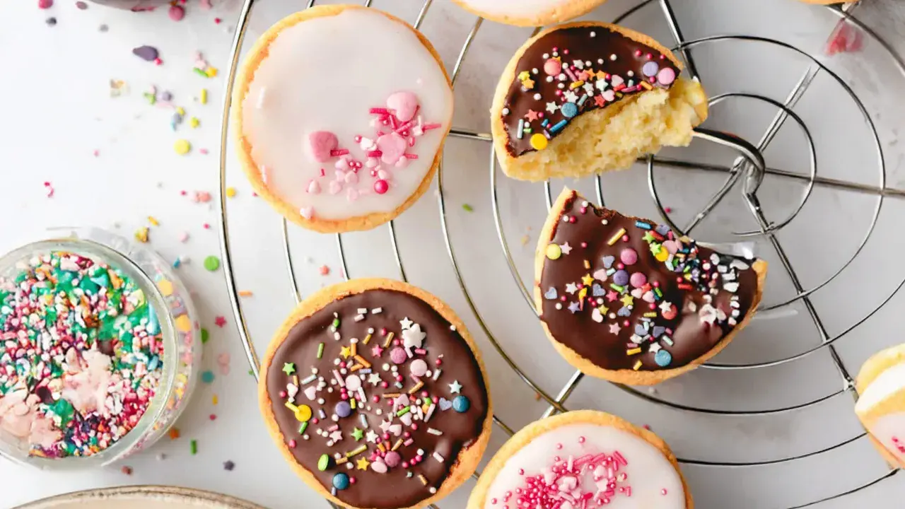 Bunte mini Amerikaner mit Schokoladen- und Zuckerguss, bestreut mit Streuseln und Herzen, auf einem Kuchengitter.