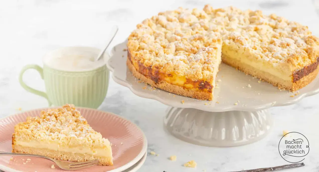 Ein saftiger Streuselkuchen mit Puddingfüllung, bestäubt mit Puderzucker. Daneben eine Tasse Kaffee und ein Stück Kuchen auf einem Teller.