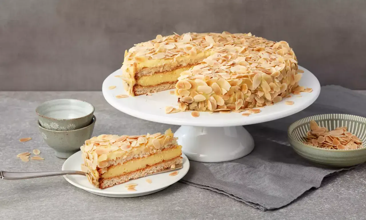 Eine köstliche schwedische Mandeltorte, üppig mit Mandelblättchen bedeckt, wird auf einem Tortenständer präsentiert. Ein Stück ist bereits auf einem Teller angerichtet.