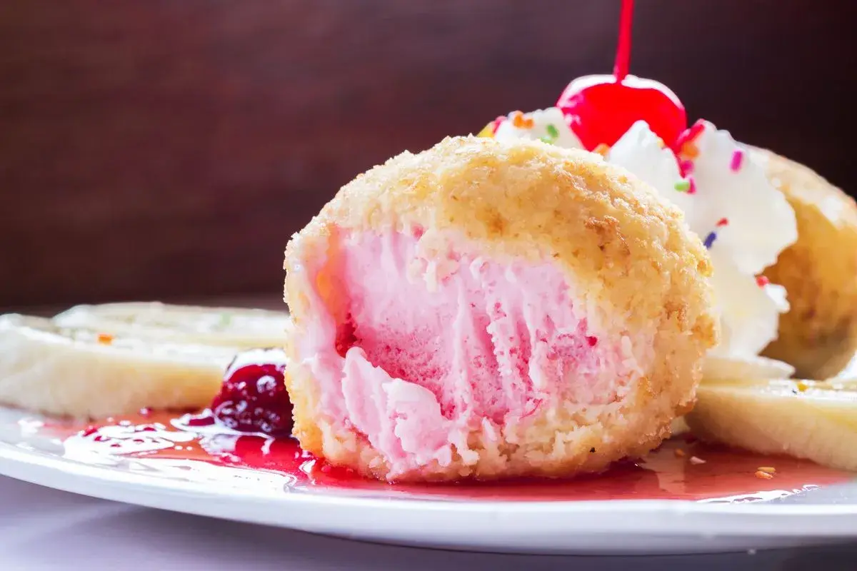 Ein knusprig gebratenes Eis mit rosa Füllung, garniert mit Sahne, Kirsche und bunten Streuseln. Dazu Bananenscheiben und rote Sauce.