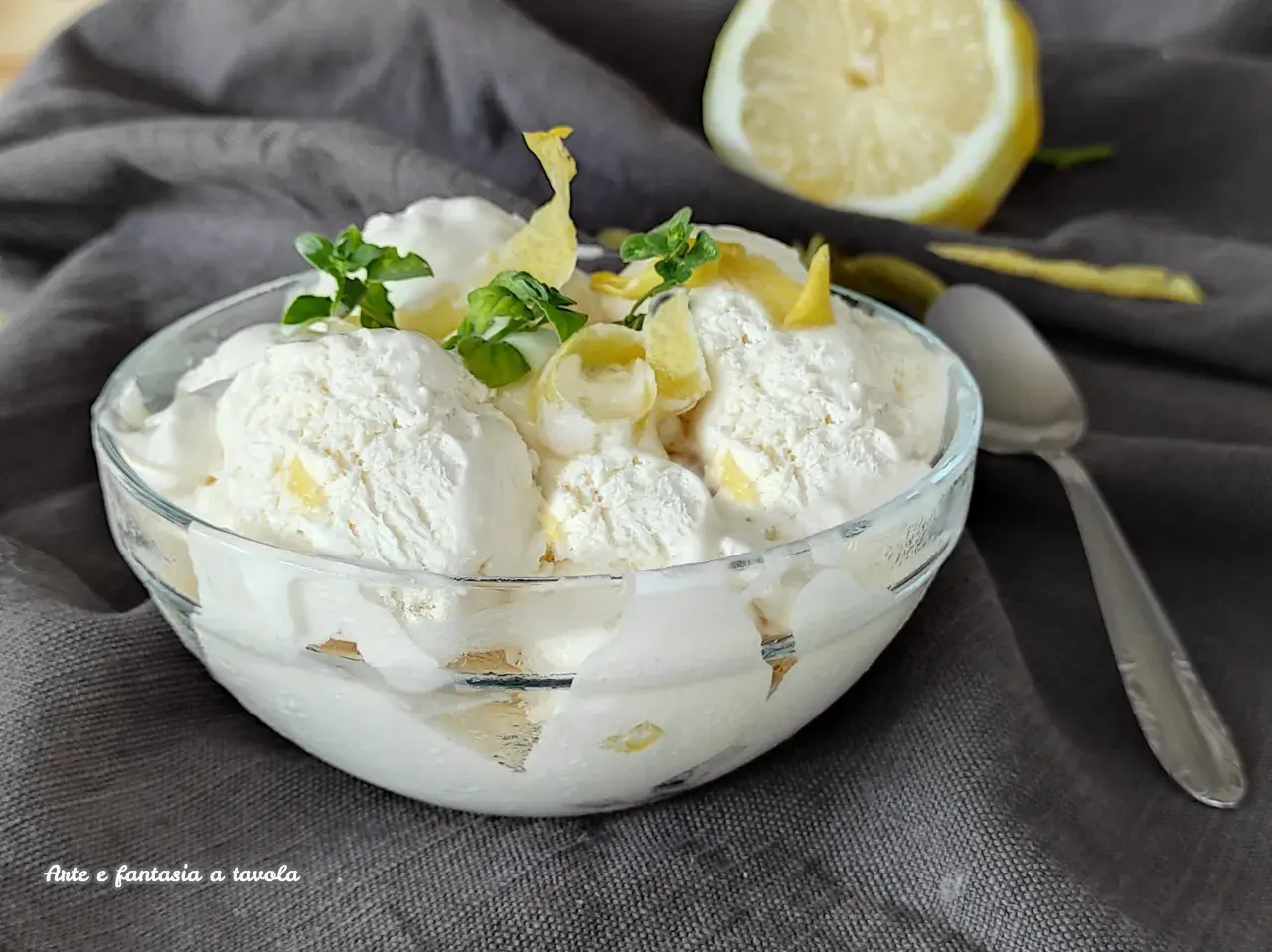Ein Glas cremiges zitronen eis, garniert mit Minze und Zitronenschale, daneben eine halbe Zitrone und ein Löffel.