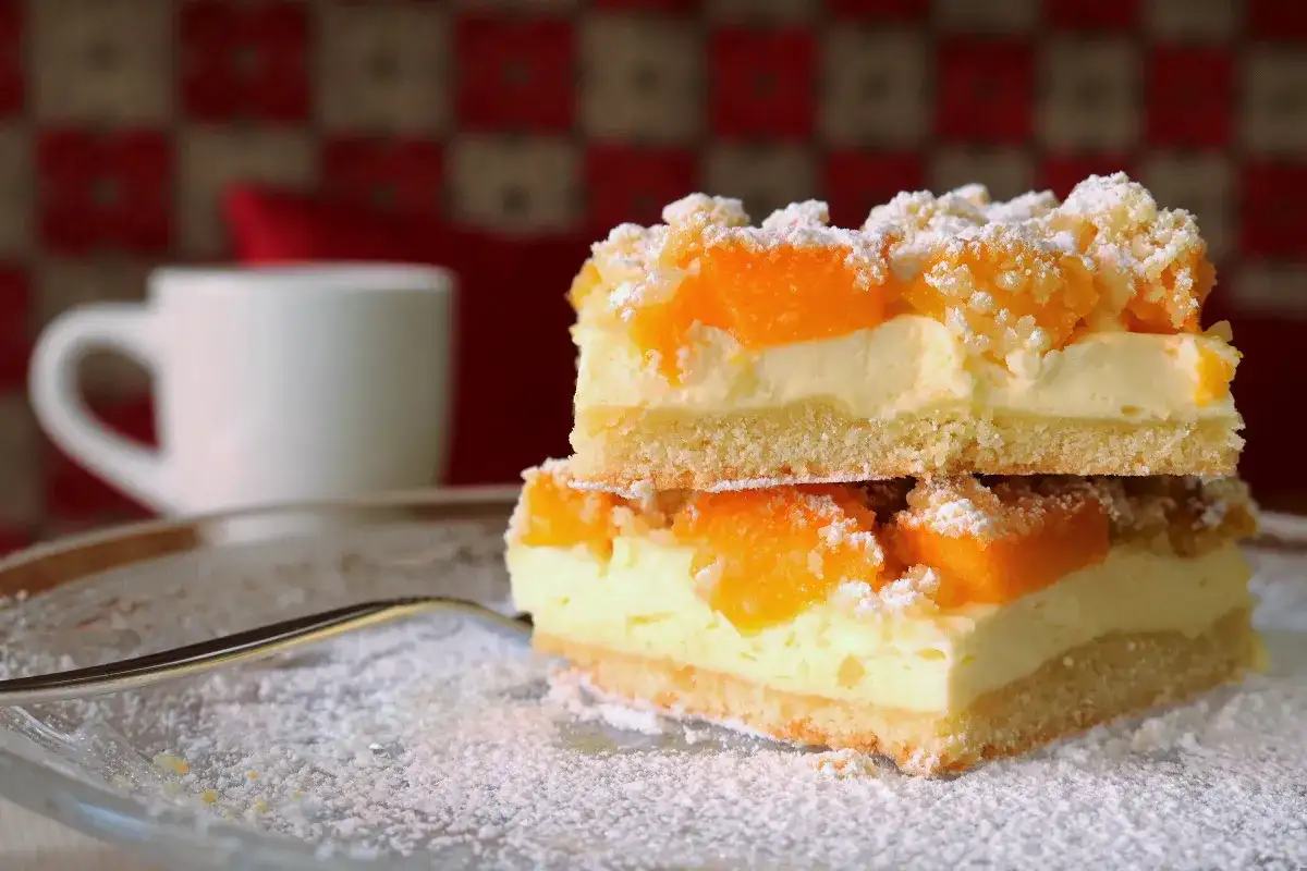 Zwei Stücke Mandarinen Schmand Kuchen Blech auf einem Teller, bestäubt mit Puderzucker. Im Hintergrund eine weiße Tasse.