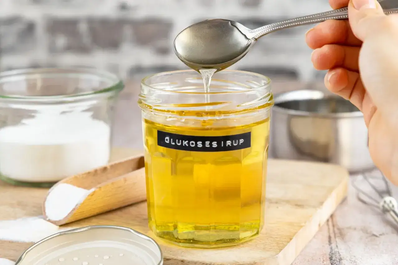 Ein Glas mit selbstgemachtem Glukosesirup, der von einem Löffel tropft. Zutaten wie Zucker sind daneben zu sehen.