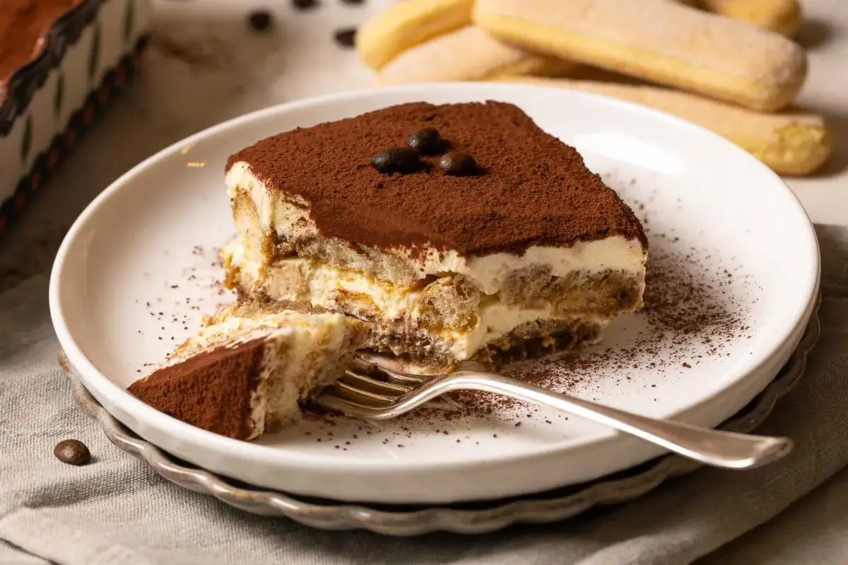 Ein Stück Tiramisu, das nach einem einfachen tiramisu rezept gemacht wurde, liegt auf einem Teller. Kakaopulver bestäubt die cremige Schicht und die Löffelbiskuits.