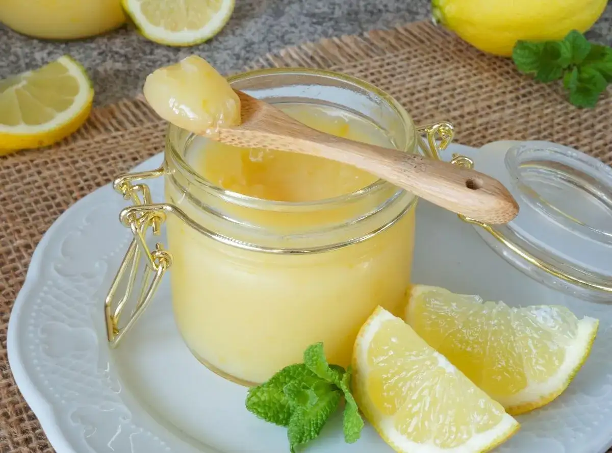Ein Glas mit cremigem Lemon Curd, garniert mit Zitronenspalten und Minze. Ein Holzlöffel mit einer Portion Lemon Curd ruht auf dem Glas.