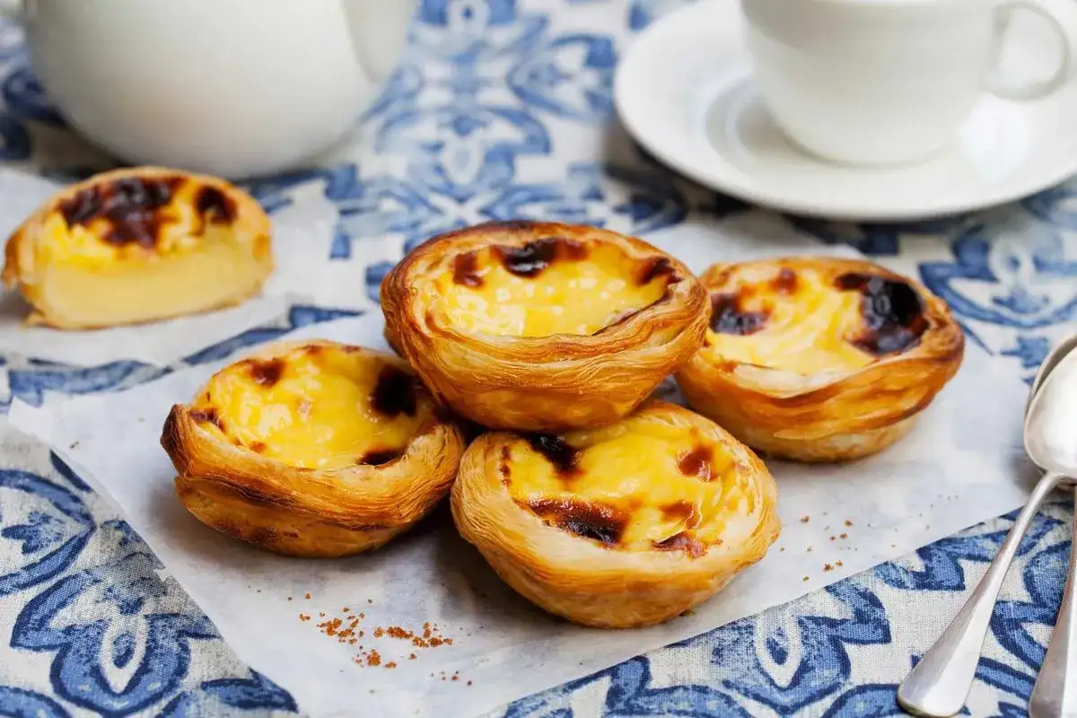 Vier goldbraune, knusprige Pastel de Nata mit cremiger Füllung und karamellisierten Spitzen, auf weißem Papier arrangiert.