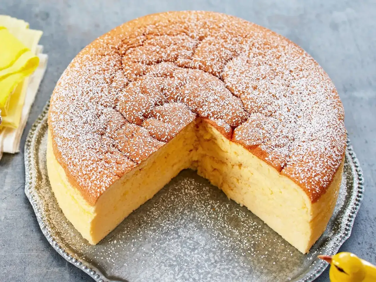 Ein fluffiger, goldbrauner low carb kuchen, bestäubt mit Puderzucker, auf einer silbernen Platte. Ein kleines gelbes Vogel-Dekoration sitzt daneben.