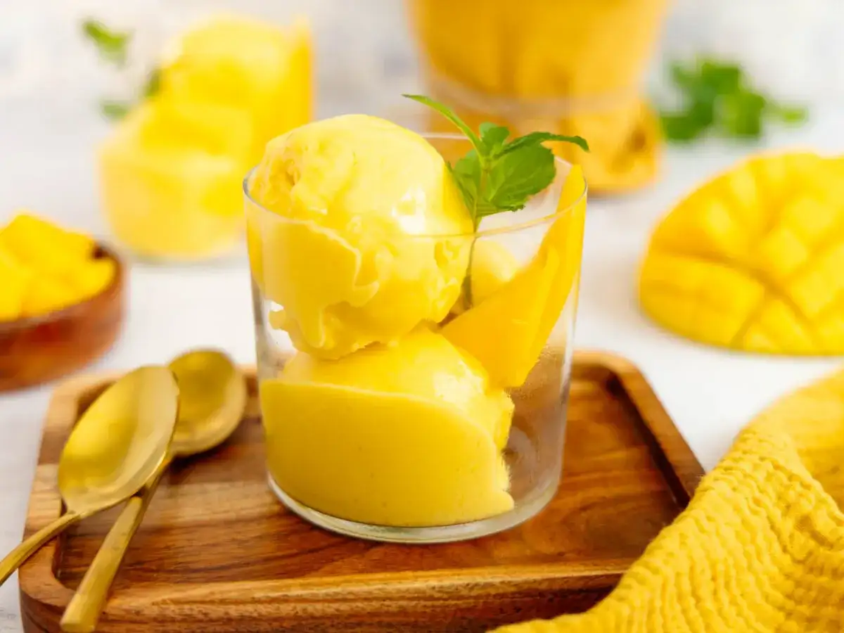 Erfrischendes Mango Eis in einem Glas, garniert mit Minze und Mangostücken. Daneben liegen goldene Löffel auf einem Holztablett.