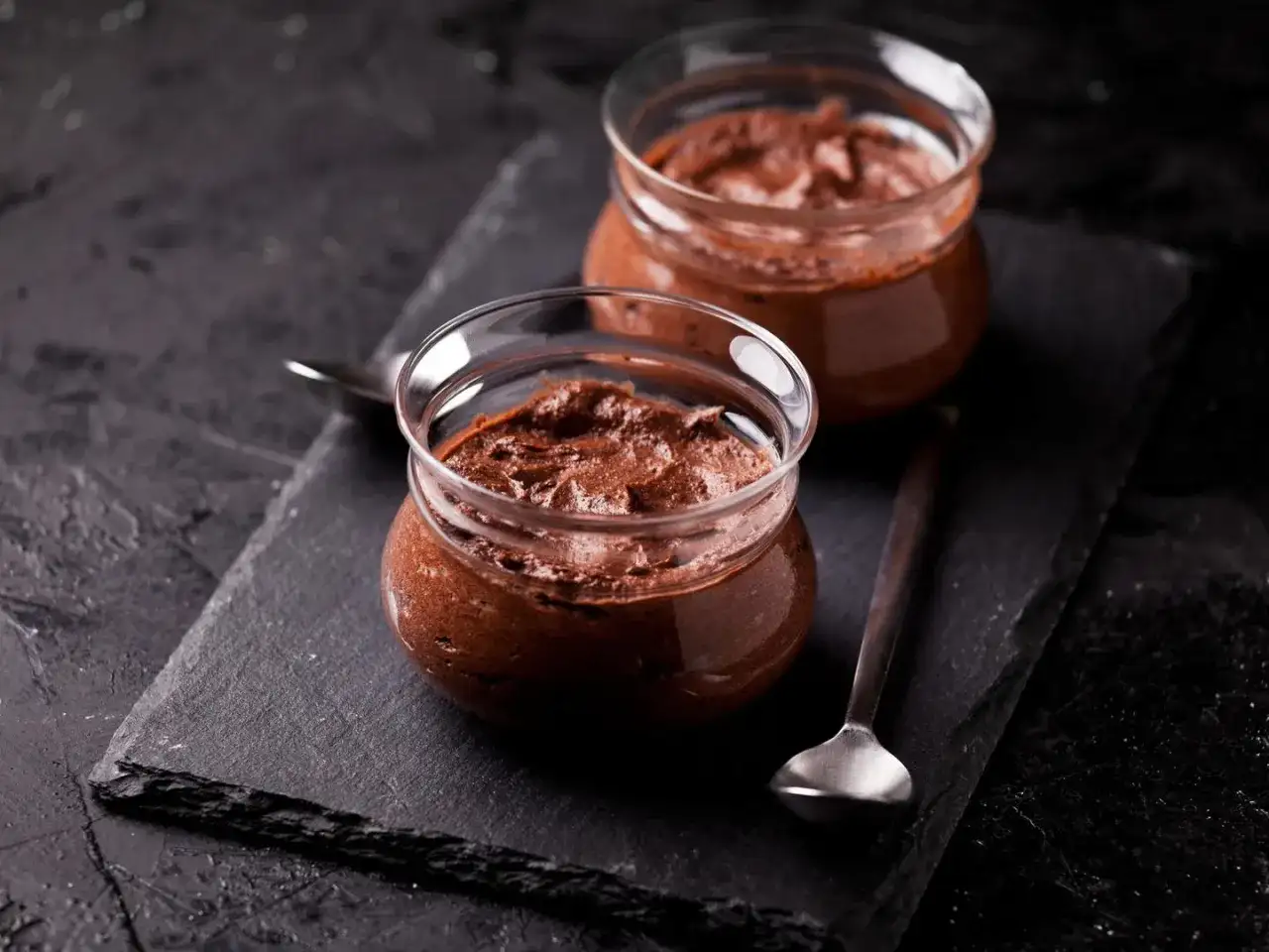 Zwei Gläser mit luftiger Mousse au Chocolat stehen auf einem dunklen Stein. Ein Löffel liegt daneben, bereit zum Genießen.
