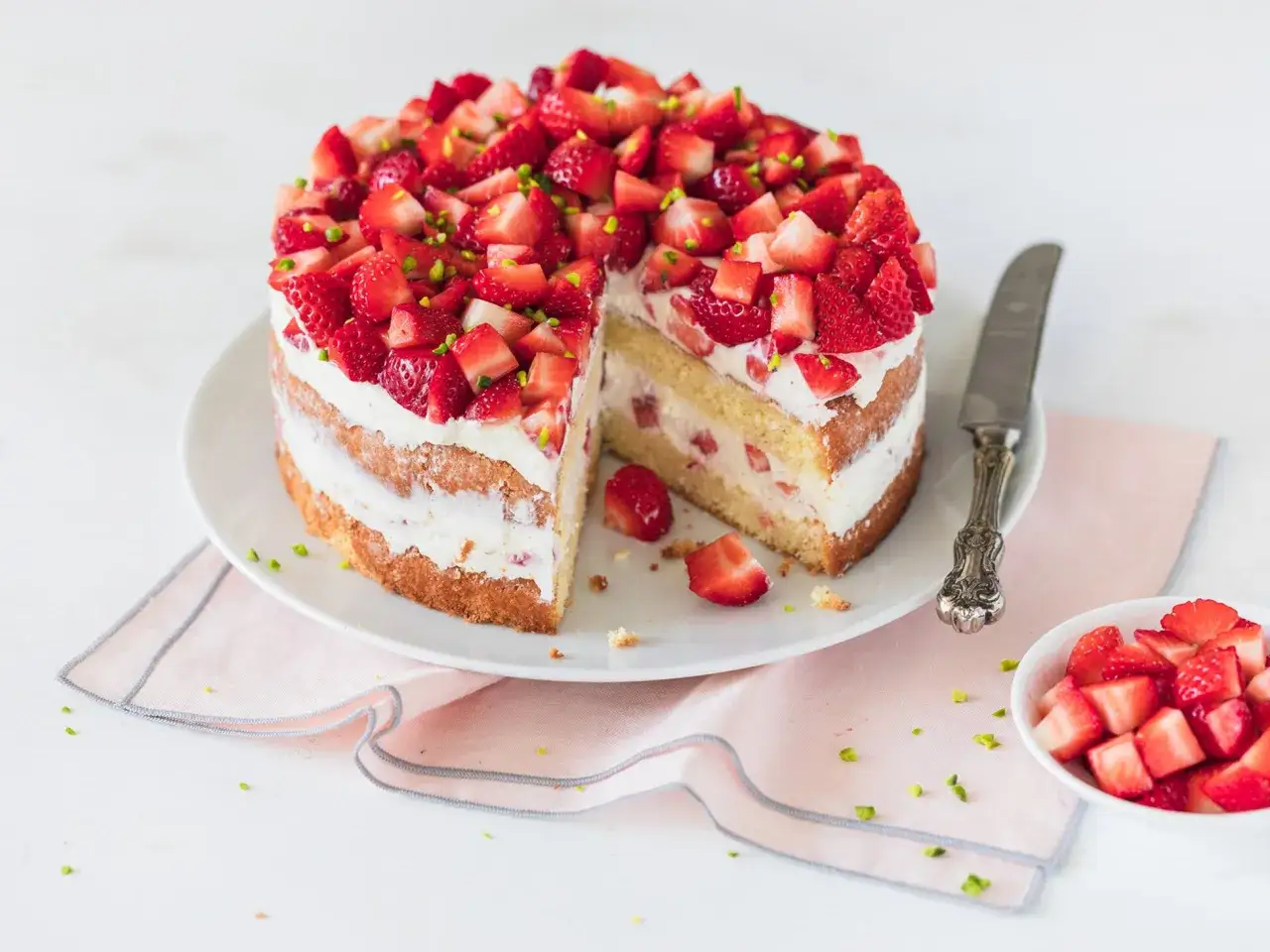 Eine köstliche Erdbeer Sahne Torte, üppig belegt mit frischen Erdbeeren und Pistazien. Ein Stück ist bereits angeschnitten.