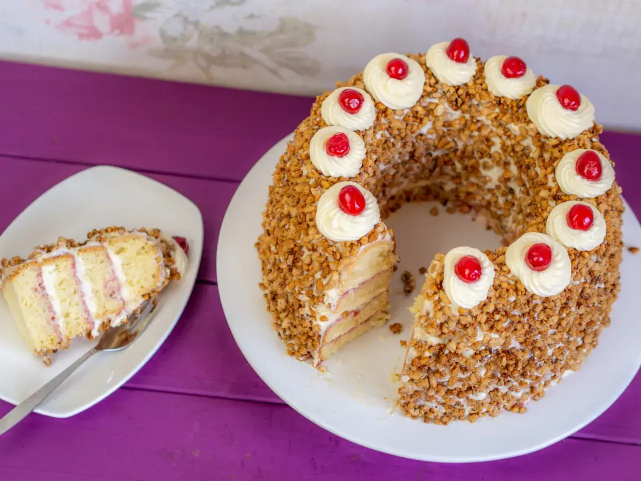 Ein köstlicher Frankfurter Kranz, umhüllt von Nüssen und verziert mit Sahnetuffs und Kirschen. Ein Stück ist bereits auf einem Teller angerichtet.