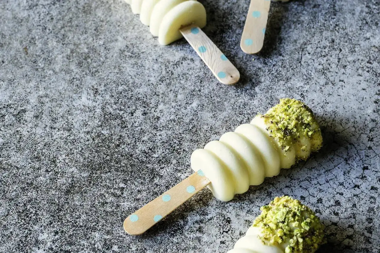 Apfeleis selber machen ohne Eismaschine: Cremiges Eis mit Pistazien-Topping auf Holzstielen mit blauen Punkten.