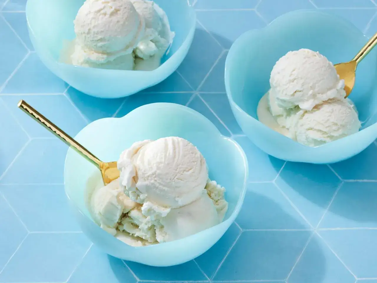 Drei Schalen mit cremigem Vanilleeis, das wie Sahne aussieht, auf einem blauen Fliesenmuster. Goldene Löffel warten darauf, das köstliche Eis zu genießen.