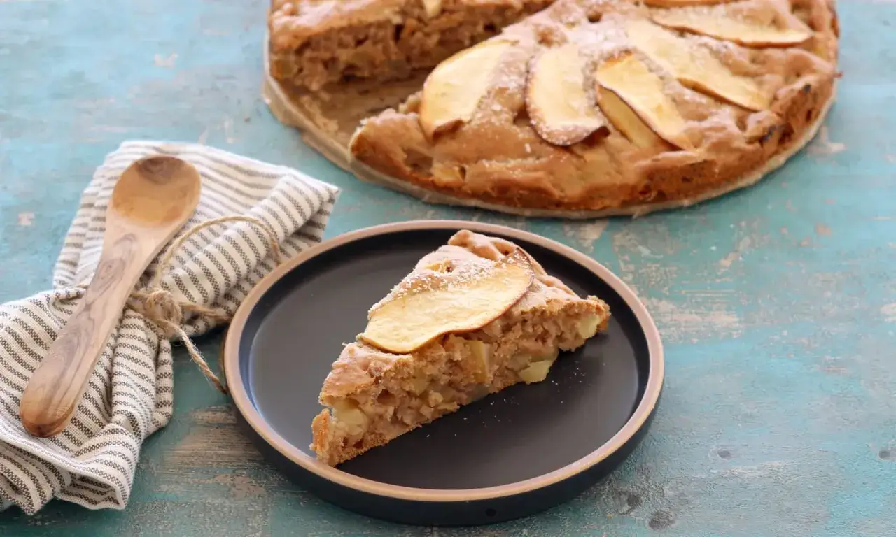 Ein Stück Apfelkuchen ohne Zucker auf einem Teller, daneben ein ganzer Kuchen, ein Holzlöffel und eine Serviette.