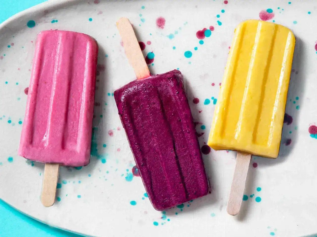 Drei bunte Fruchteis am Stiel: rosa, lila und gelb, auf einer weißen Platte mit Farbspritzern.