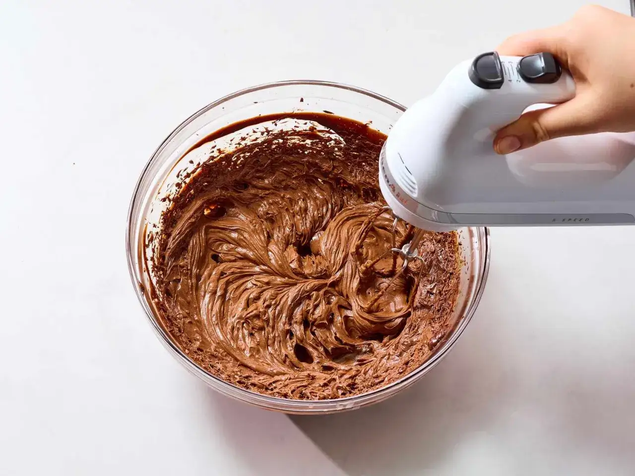 Eine Hand mixt mit einem Handmixer eine cremige Schokoladenmasse in einer Glasschüssel. Perfekt für ein einfaches ganache rezept.