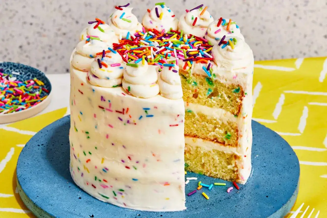Ein bunter Konfetti Kuchen mit weißer Glasur und Streuseln. Er ist aufgeschnitten und zeigt mehrere Schichten.