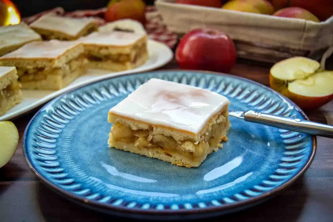 Ein Stück saftiger Apfelkuchen mit Zuckerguss auf blauem Teller. Daneben liegen Äpfel und weitere Kuchenstücke.