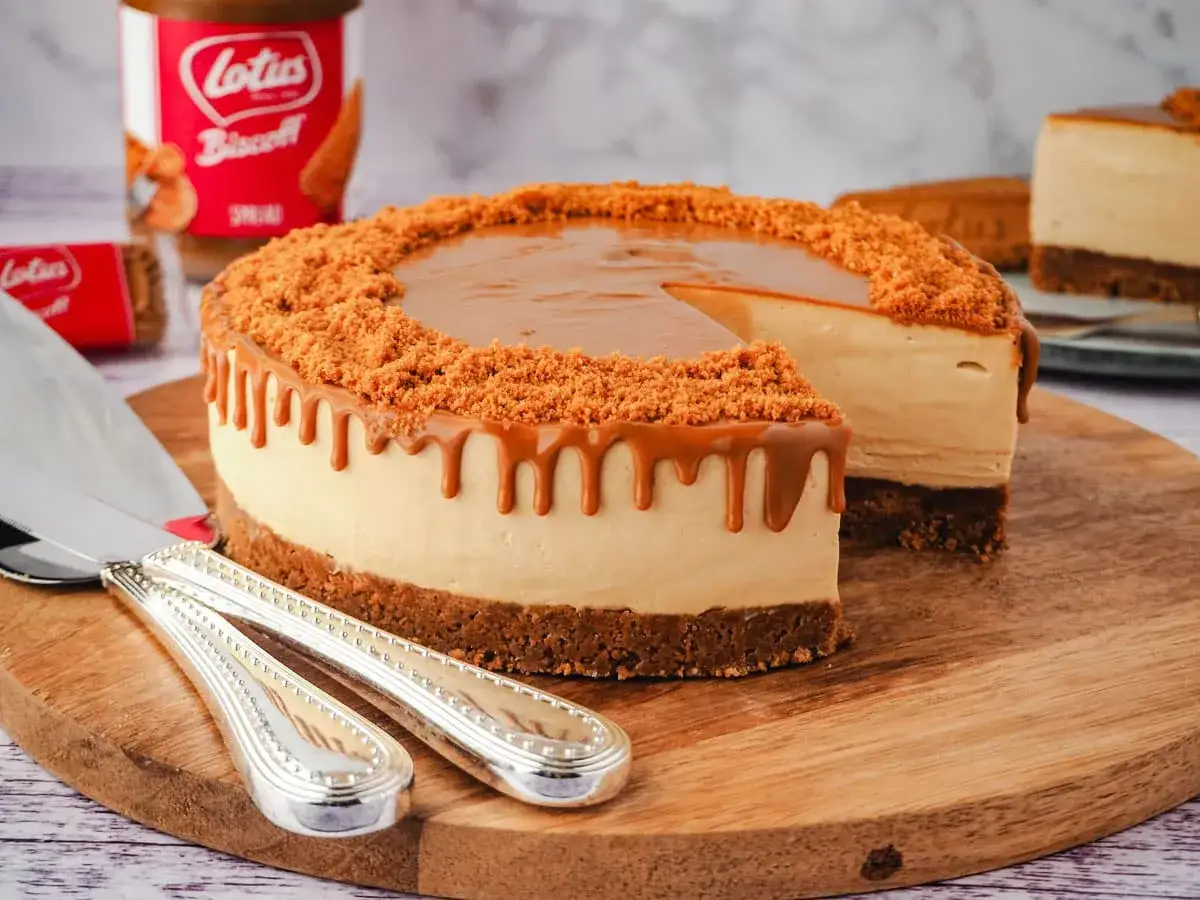 Ein köstlicher Lotus Cheesecake mit Karamell-Drip und Keksbröseln. Daneben ein Glas Lotus Biscoff Spread.