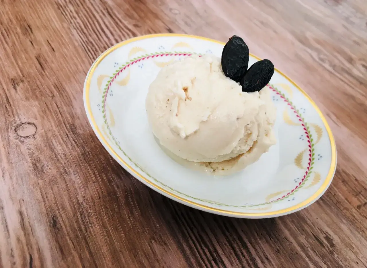 Ein Löffel cremiges Tonka Eis auf einer verzierten Schale, garniert mit zwei dunklen Rosinen.