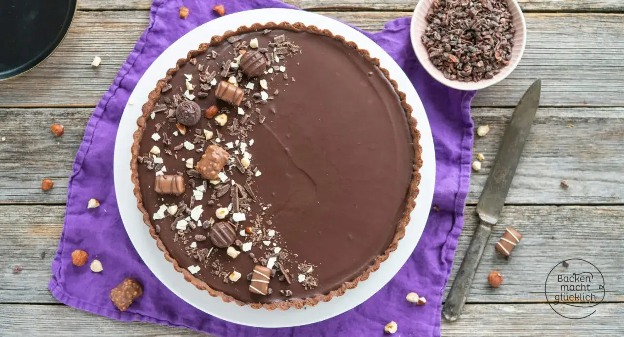Eine köstliche Schoko Tarte, verziert mit Schokostückchen, Nüssen und Pralinen, liegt auf einem lila Tuch.