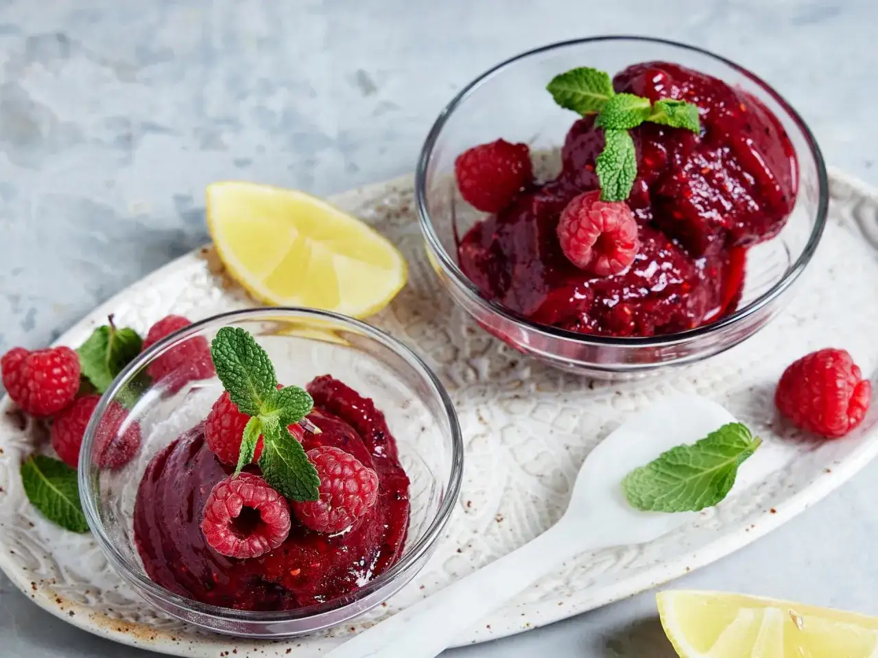 Zwei Schalen mit erfrischendem Himbeer-Sorbet-Eis, garniert mit frischen Himbeeren und Minze, daneben Zitronenspalten.