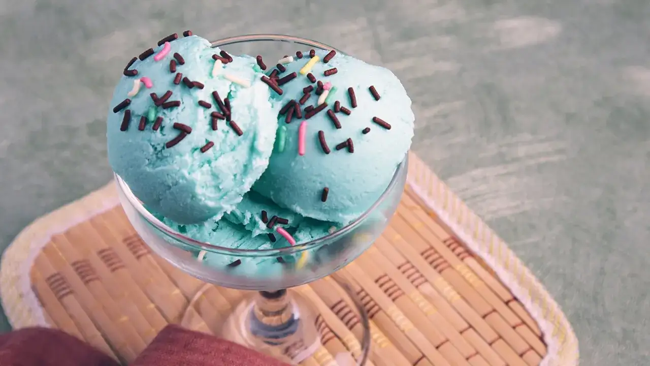 Ein Glas mit engelblauem Eis, bestreut mit Schokostreuseln und bunten Zuckerperlen, steht auf einem Bambusteller.