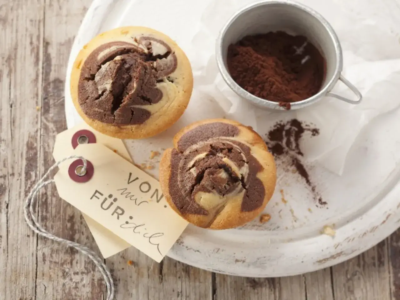 Zwei zupfkuchen muffins mit Marmormuster, daneben ein kleines Töpfchen mit Kakaopulver und ein Anhänger mit der Aufschrift 