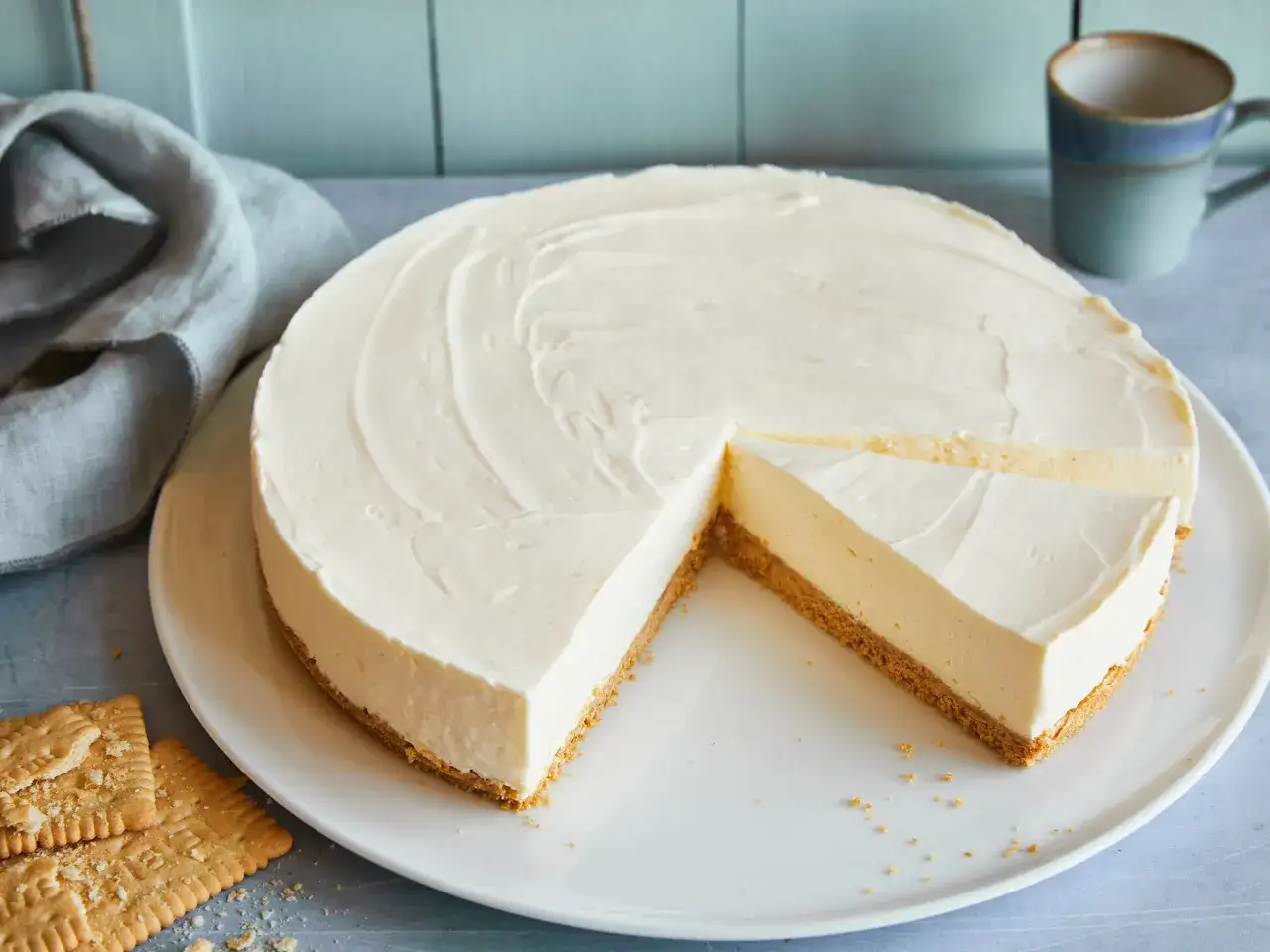 Cremiger cheesecake ohne backen auf einem Teller, mit einem Stück herausgeschnitten. Daneben liegen Kekse und eine Tasse.