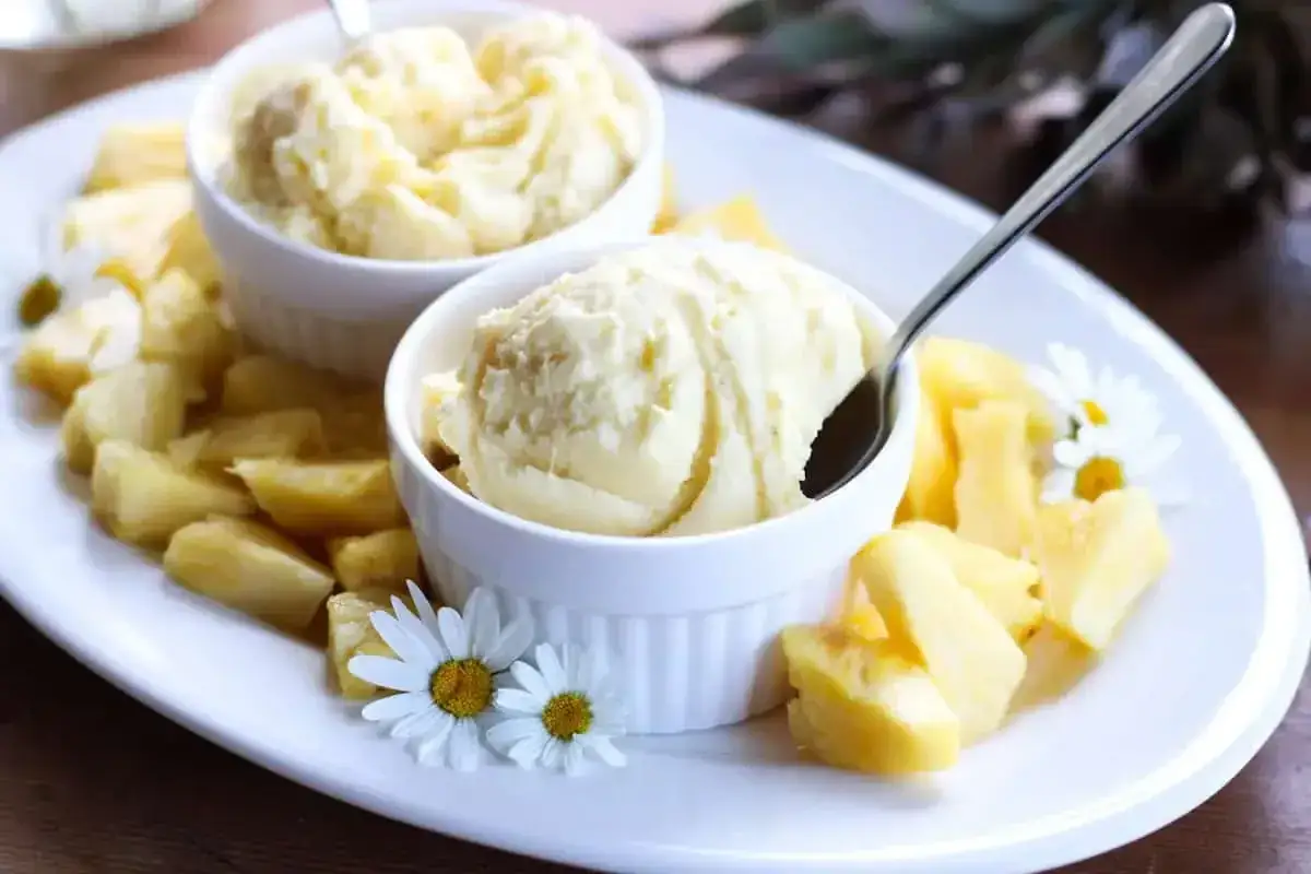 Zwei Schalen cremiges Ananas Eis, umgeben von frischen Ananasstücken und Gänseblümchen, auf einem weißen Teller.