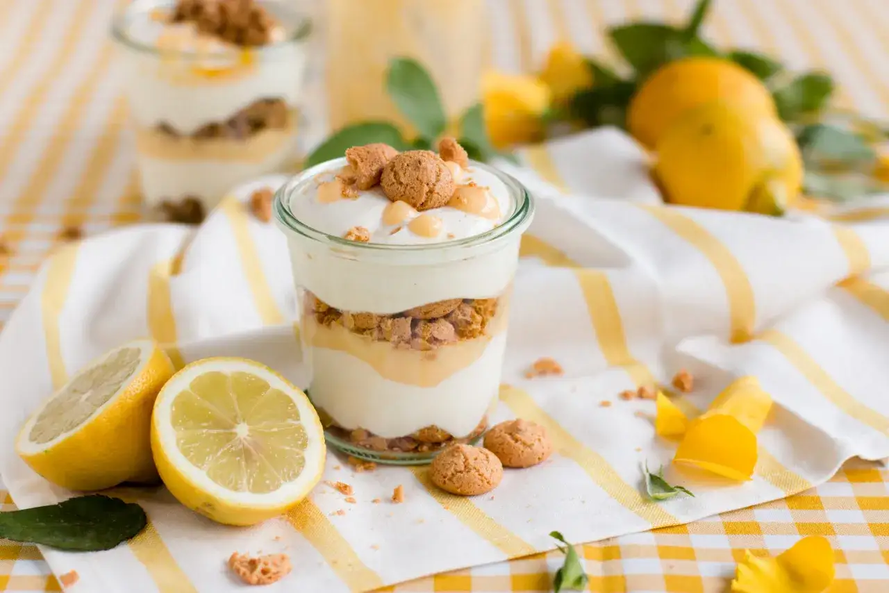 Ein Glas mit cremigem Zitronen Tiramisu, garniert mit Keksen und Zitronenspalten daneben.