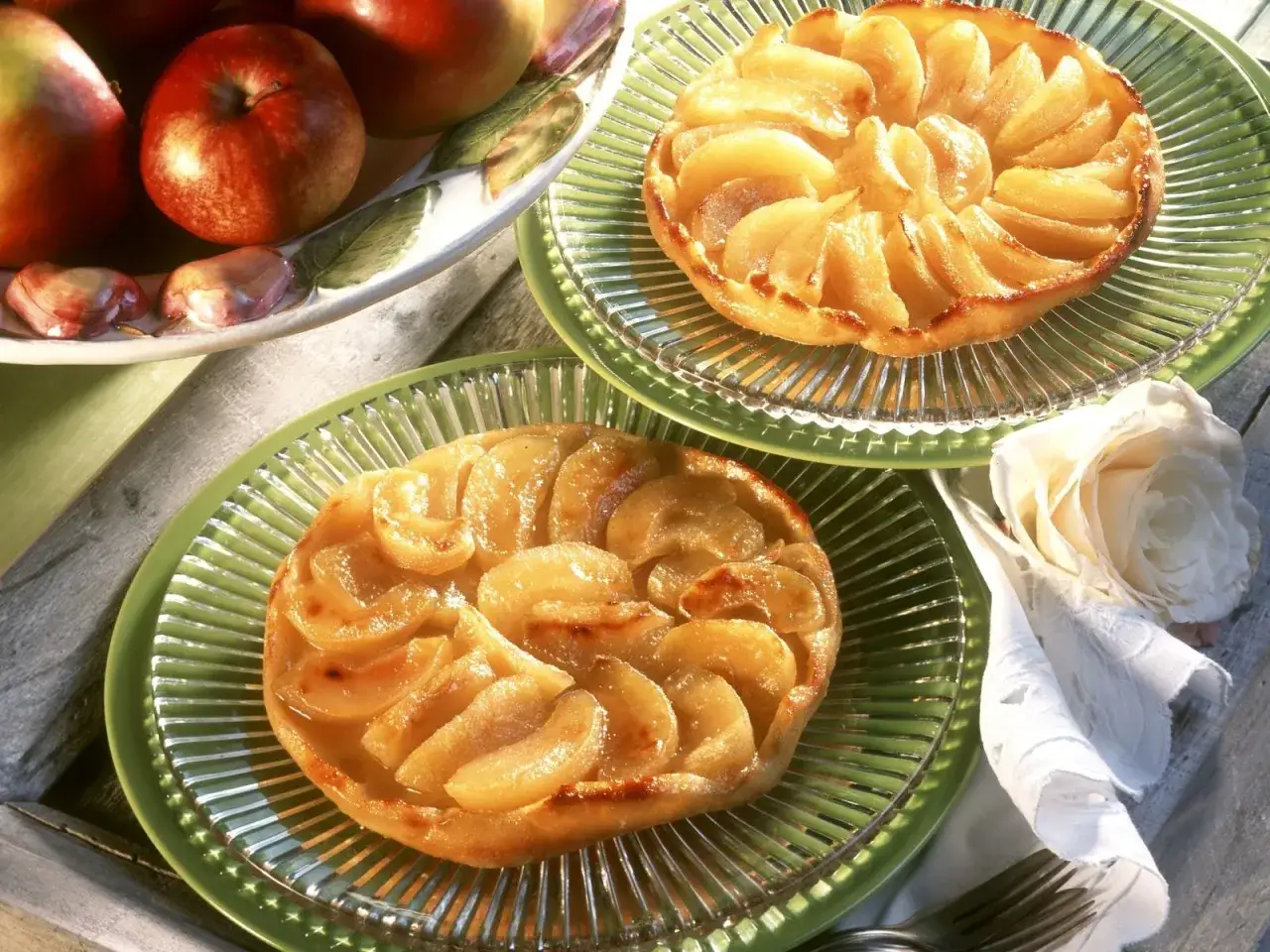 Zwei goldbraune Tarte Tatin mit karamellisierten Äpfeln auf grünen Tellern, daneben frische Äpfel und eine weiße Rose.