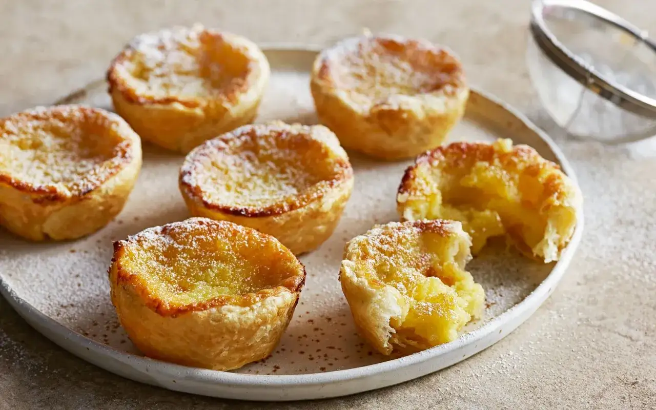 Sechs goldbraune, mit Puderzucker bestäubte Pastel de Nata auf einem Teller. Eines ist angeknabbert und zeigt die cremige Füllung.