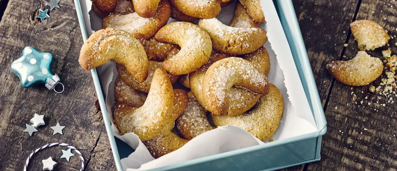 Vanillekipferl, zart und mit Puderzucker bestäubt, liegen in einer hellblauen Dose. Weihnachtliche Sterne und eine Kugel schmücken den Holztisch.