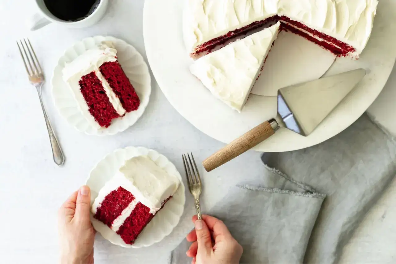 Ein Stück saftiger red velvet Kuchen mit cremiger Glasur. Daneben ein ganzer Kuchen, zwei Teller mit je einem Stück und eine Kaffeetasse.